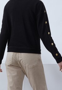 Pullover nero texturizzato con bottoni dorati lungo le maniche, abbinato a jeans beige chiari con tasche posteriori e una vestibilità aderente.