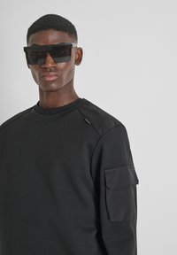 Felpa nera con scollatura tonda, dettagli texturizzati sulle spalle e taschino sulla manica. Il modello indossa occhiali da sole neri oversize.