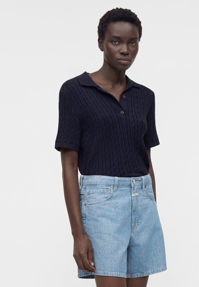 Γυναίκα που φοράει μπλε-σκούρο πουλόβερ με κοντά μανίκια και ανοιχτόχρωμο τζιν σορτς, στέκεται μπροστά σε απλό ανοιχτό φόντο.