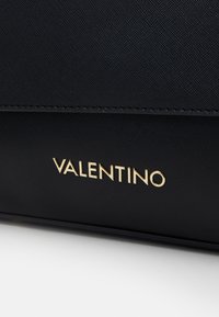 A bolsa texturizada preta apresenta um logotipo dourado "VALENTINO". O design é elegante, com um acabamento suave e sutis detalhes em costura.