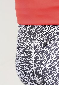 Černobílé legíny s vzorováním a vysokým pasem, které mají strukturovaný design. Na zadní straně je viditelná malá zipová kapsa.