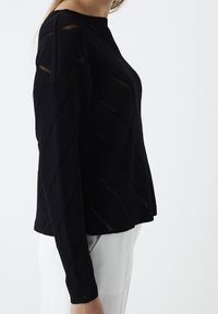 Czarny sweter wykonany z dzianiny z ukośnymi wycięciami, okrągłym dekoltem i długimi rękawami. Prezencja jest luźna, z opadającą sylwetką.