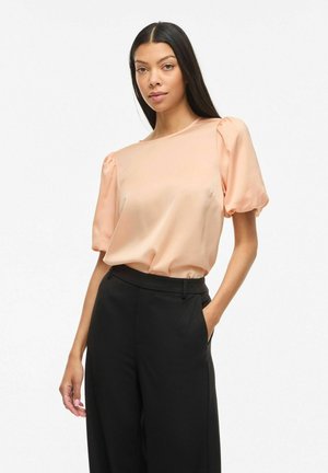 Femme aux longs cheveux noirs portant un chemisier en satin pêche à manches bouffantes et un pantalon noir taille haute, posant avec une main dans la poche.