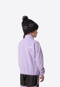 Jasnofioletowa polarowa kurtka z wysokim kołnierzem, z białym logo. Dziecko nosi czarną czapkę z dzianiny z wzorem i pomponem.