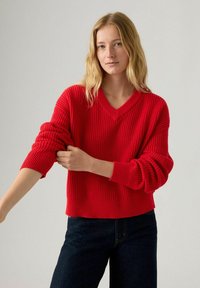 Czerwony sweter z prążkowanego dzianiny z dekoltem w serek i luźnym krojem, z grubymi, podwiniętymi rękawami i lekko skróconym dołem. W zestawie z ciemnymi dżinsami.