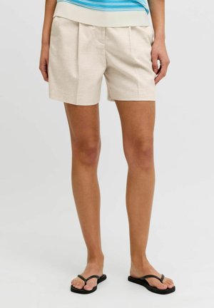 Pantalones cortos beige plisados combinados con una camiseta de rayas azul y blanca y chanclas negras sobre piernas desnudas con un fondo liso.