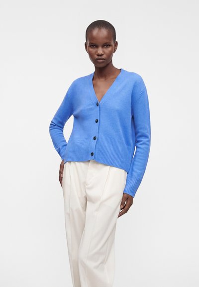 Femme portant un cardigan bleu à col en V boutonné et un pantalon blanc ample, debout avec une main dans la poche sur un fond uni.