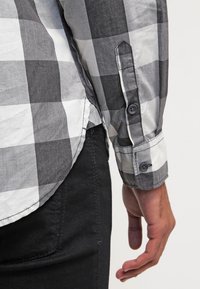 Camicia a maniche lunghe grigia e bianca a quadri con polsini abbottonati, realizzata in tessuto leggero, con orlo curvo e cuciture dettagliate.