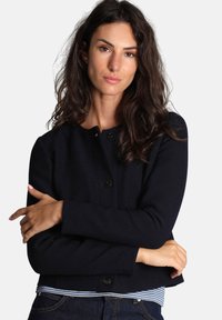 Giacca struttura in blu navy, con scollo rotondo, chiusura a bottoni e maniche lunghe, indossata sopra una camicia a righe.