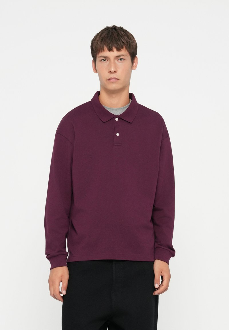 Long-sleeve polo shirt in deep plum color with a classic collar, three-button placket, and ribbed cuff details. Soft, textured fabric.