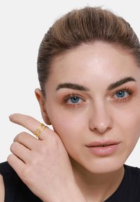 Goldfarbener, verdrehter Ring am Zeigefinger, mit einem strukturierten Design und verschlungenen Bändern, vor einem hellen Hautton und einem schwarzen Oberteil.