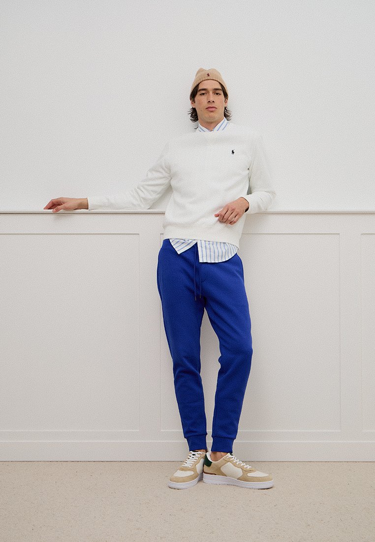 Biała sweter na niebieską koszulę w paski, zestawiona z niebieskimi joggerami i beżowymi trampkami. Modelka ma na sobie jasnobeżową czapkę beanie.