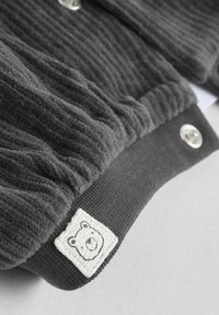 Halli ribadega kardigan, mis on valmistatud pehmest kangast ja millel on hõbedased nööbid. Omab musta kudumisega mansetti ja valget karu logo patši ääres.