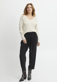 Pull en maille beige à col en V, associé à un pantalon noir taille haute avec une ceinture nouée, et des bottines noires à motifs.