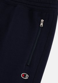Marineblauwe sweatpants van zacht materiaal hebben een zijzak met rits en een klein geborduurd logo. Elastische tailleband met een gestructureerd stiksel.