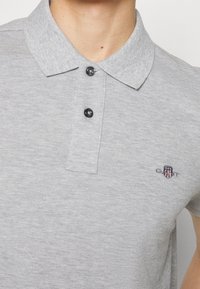 Polo gris clair avec deux boutons noirs et un petit logo brodé en forme de bouclier sur la poitrine gauche, porté par une personne.