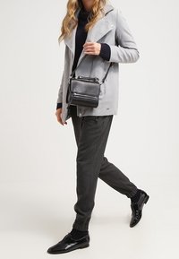 Šedý kabát s klopovým límcem, černý rolák, tmavě šedé zúžené kalhoty a lesklé černé boty; nese malou černou crossbody tašku.