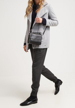 Šedý kabát s klopovým límcem, černý rolák, tmavě šedé zúžené kalhoty a lesklé černé boty; nese malou černou crossbody tašku.