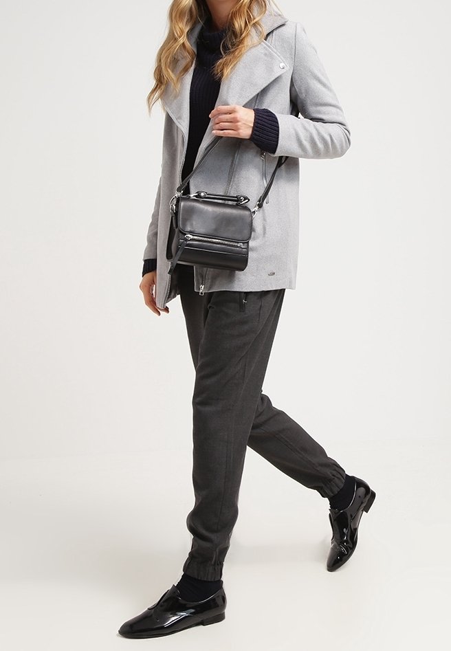 Šedý kabát s klopovým límcem, černý rolák, tmavě šedé zúžené kalhoty a lesklé černé boty; nese malou černou crossbody tašku.