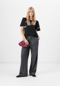 Femme portant un haut noir avec des bordures blanches, un pantalon large gris, des chaussures bordeaux et tenant un sac à main bordeaux avec une bandoulière.