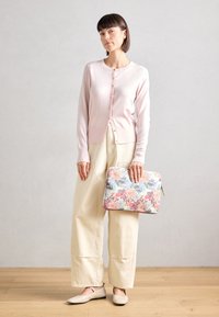 Cardigan rose clair avec des boutons, pantalon large crème et ballerines beiges. Tenant un sac rembourré à motifs floraux dans des couleurs douces.