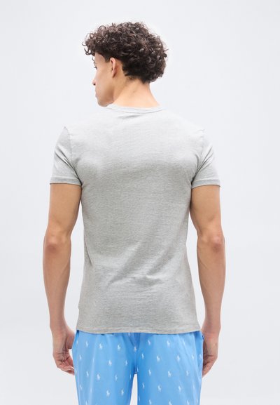 Νέος άνδρας με σγουρά μαλλιά στέκεται γυρισμένος με την πλάτη, φοράει ανοιχτό γκρι κοντομάνικη μπλούζα και μπλε σορτς με λευκά λογότυπα.