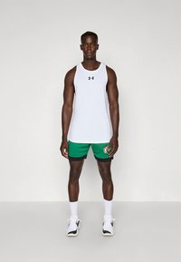 Vitt linne och gröna shorts med svarta detaljer och en logotyp. Modellen står i sportskor och visar upp en passform och ett andningsbart tyg.