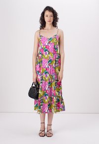 Blumiges Maxikleid mit lebendigen rosa und grünen Designs, dünnen Spaghettiträgern und lockerer Passform. Das Model hält eine kleine schwarze Handtasche.
