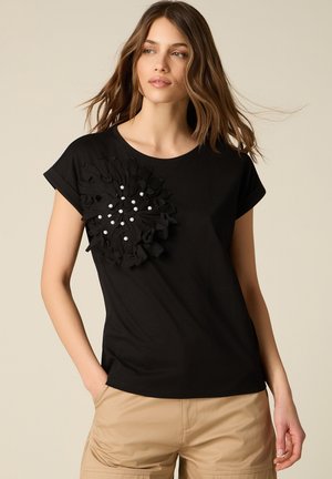 Donna che indossa una maglietta nera a maniche corte con dettagli floreali arricciati e accenti di perle sul petto, abbinata a pantaloni beige, in piedi contro uno sfondo semplice.