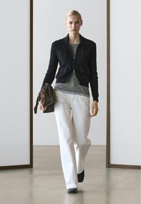 Donna che cammina al chiuso indossando un maglione nero con cerniera, camicia grigia, pantaloni bianchi, scarpe nere e tiene una borsa marrone nella mano sinistra.