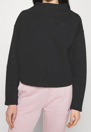 Femme portant un sweat-shirt noir à manches longues Nike et un pantalon de survêtement rose clair, debout les bras détendus le long du corps.