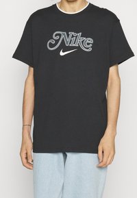 Svart bomullströja med en stor broderad "Nike"-logotyp i grått och vitt, rund halsringning och korta ärmar.