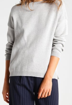 Light gray sweater made of soft fabric, featuring a round neck, dropped shoulders, and ribbed cuffs, paired with navy pinstriped trousers.