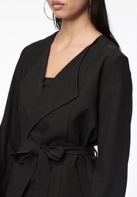 Manteau portefeuille noir en tissu texturé avec un col cranté, des manches longues et une ceinture nouée à la taille.