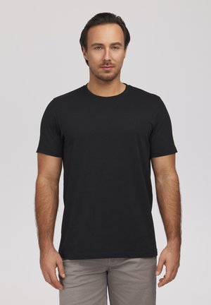 Mężczyzna o ciemnych włosach, ubrany w jednolitą czarną koszulkę z krótkim rękawem i jasnoszare spodnie, stojący na tle jasnoszarego tła.