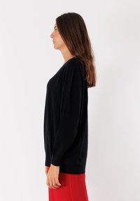 Donna con lunghi capelli castani in profilo, indossa un maglione nero a maniche lunghe e pantaloni rossi su uno sfondo bianco.