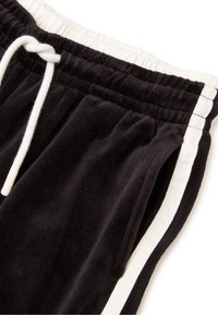 Pantalones de chándal de terciopelo negro con cinturilla elástica, cordón blanco y rayas laterales en contraste en blanco. Textura suave, corte relajado.