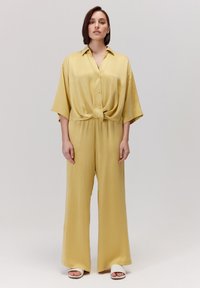 Żółty satynowy kombinezon z węzłem z przodu, krótkimi rękawami i szerokimi nogawkami. W zestawieniu z białymi butami na otwartym palcu na neutralnym tle.