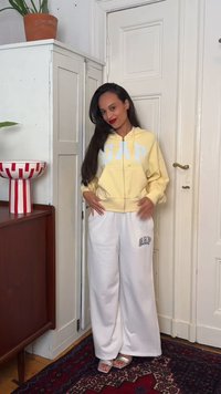 Γυναίκα με μακριά σκουρόχρωμα μαλλιά, που φοράει κίτρινο φούτερ GAP με κουκούλα και λευκό παντελόνι GAP με φαρδιά μπατζάκια, στέκεται μέσα σε εσωτερικό χώρο κοντά σε μια λευκή πόρτα και ξύλινη ντουλάπα.