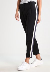 Black pants with a high waist and tapered legs, featuring a white and navy side stripe. Made from a smooth material. Worn with white sneakers.