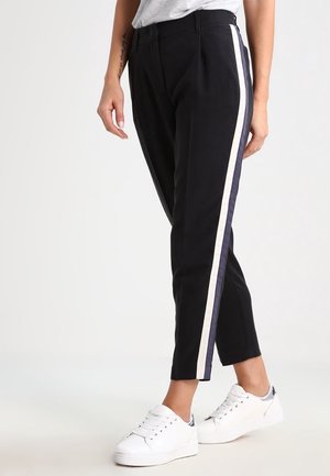 Pantalons noirs à taille haute et jambes fuselées, avec une bande latérale blanche et marine. Fabriqués à partir d'un matériau lisse. Portés avec des baskets blanches.