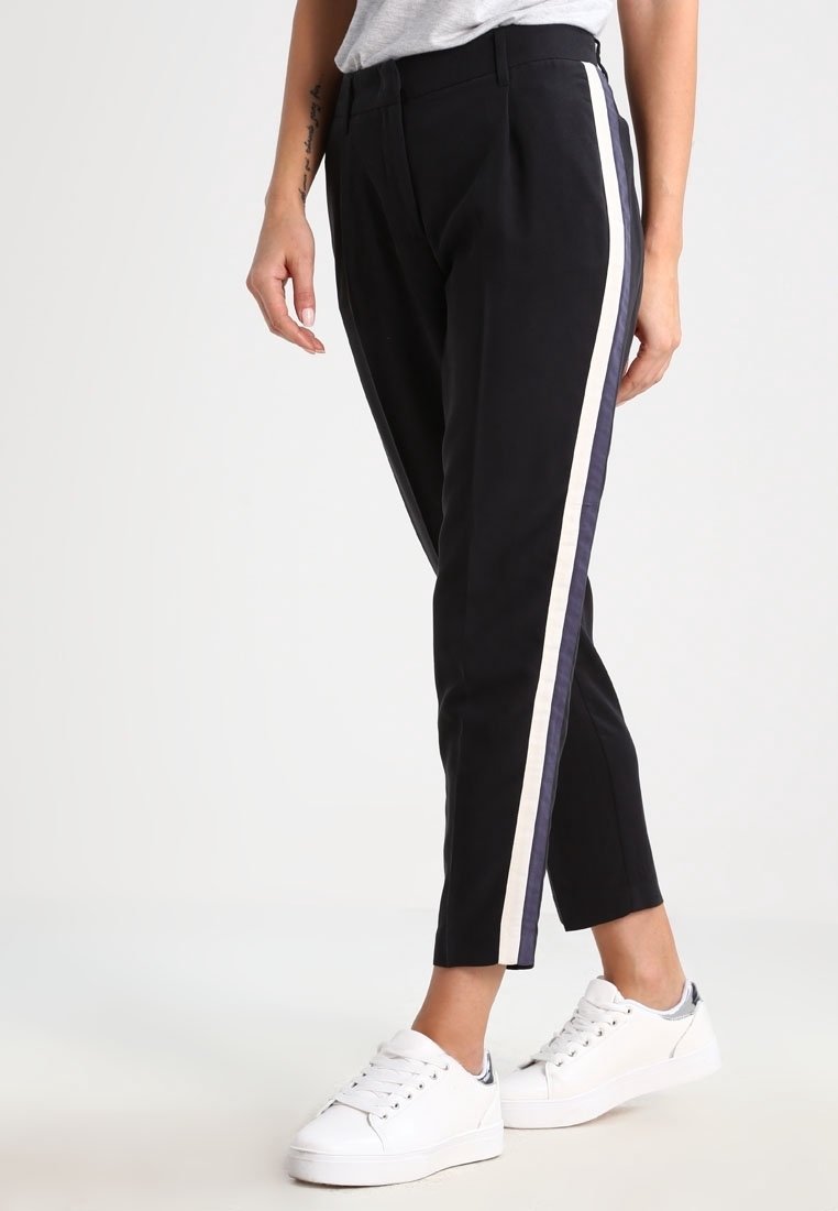 Black pants with a high waist and tapered legs, featuring a white and navy side stripe. Made from a smooth material. Worn with white sneakers.