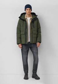 Donkergroene gewatteerde jas met capuchon, voorzien van meerdere zakken en een ritssluiting. Draagt over een beige overhemd, gecombineerd met donkere spijkerbroek en laarzen.