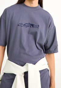 Camiseta corta de manga corta color púrpura con cuello alto, que presenta una textura suave y un gráfico de coche en azul oscuro. Se lleva una tela blanca atada a la cintura.