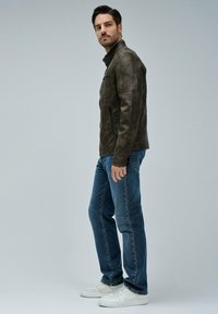 Giubbotto in pelle marrone con colletto rialzato, cerniere ai lati e una texture leggermente usurata, abbinato a jeans di denim blu e sneakers bianche.