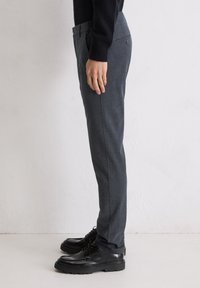 Dunkelgraue, maßgeschneiderte Hose mit strukturiertem Muster, schlanker Passform, seitlichen Taschen und einem Knopfverschluss am Rücken, getragen mit schwarzen Schuhen.
