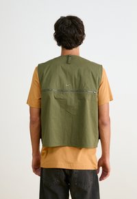 Ελαιόχρωμο αμάνικο γιλέκο με τσέπη με φερμουάρ στην πλάτη και μικρή λεπτομέρεια λογότυπου. Φορεμένο πάνω από ένα μπεζ T-shirt, επιδεικνύει χαλαρή εφαρμογή.