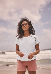 Kvinna med lockigt hår som bär solglasögon, vit kortärmad topp och rosa shorts står på en strand med vågor och molnig himmel i bakgrunden.