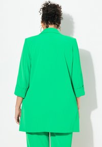 Žena s kudrnatými vlasy, oblečená v zeleném saku a odpovídajících kalhotách, stojí čelem ke bílé zdi s uvolněnýma rukama podél těla.