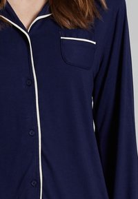 Navy blue shirt with long sleeves, featuring a pocket and white piping along the collar and button line. Smooth fabric texture.